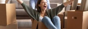 A woman sits in an empty box in a new apartment while a man pushes her from behind