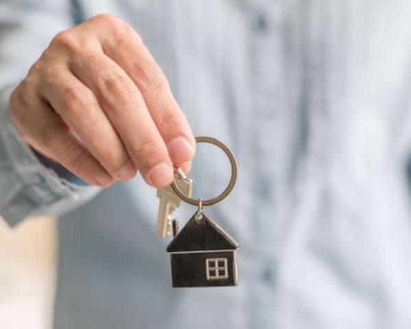 A man in a blue shirt holds out a key that's attached to a ring with a small house on it